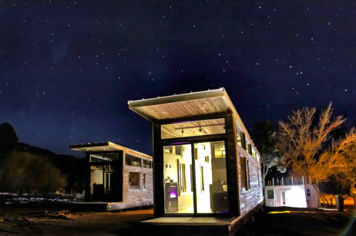 A modern cabin illuminated at night, surrounded by stars and trees in a serene outdoor setting.